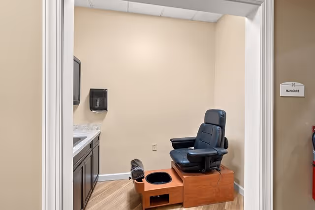 Interior view of a manicure room with a black leather manicure chair on a wooden platform, a sink with countertop and cabinets on the left, and a paper towel dispenser mounted on the wall. The room has beige walls and wood flooring. A sign outside the door reads 'MANICURE'.