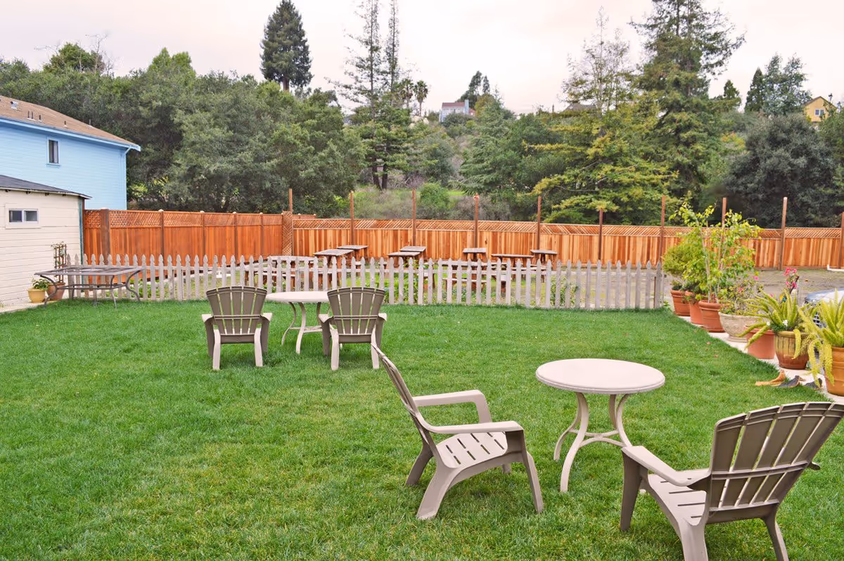 Grassy outdoor courtyard with plastic chairs and small tables, potted plants, a picket fence, and picnic benches by a wooden fence.
