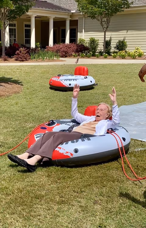 An elderly woman is sitting in an inflatable tube on a grassy lawn outside a senior living facility. She has her arms raised and appears to be enjoying herself. Another similar inflatable tube is visible in the background near the building with columns and landscaping.