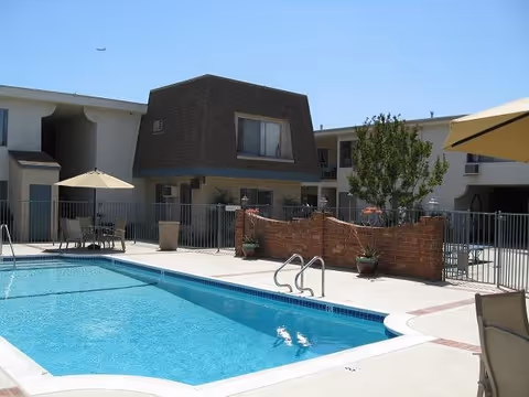 Outdoor swimming pool area at a senior living facility with clear blue water, surrounded by a concrete deck with patio tables, chairs, and umbrellas. The background shows a two-story building with windows and balconies, along with some potted plants and a tree near a brick wall.