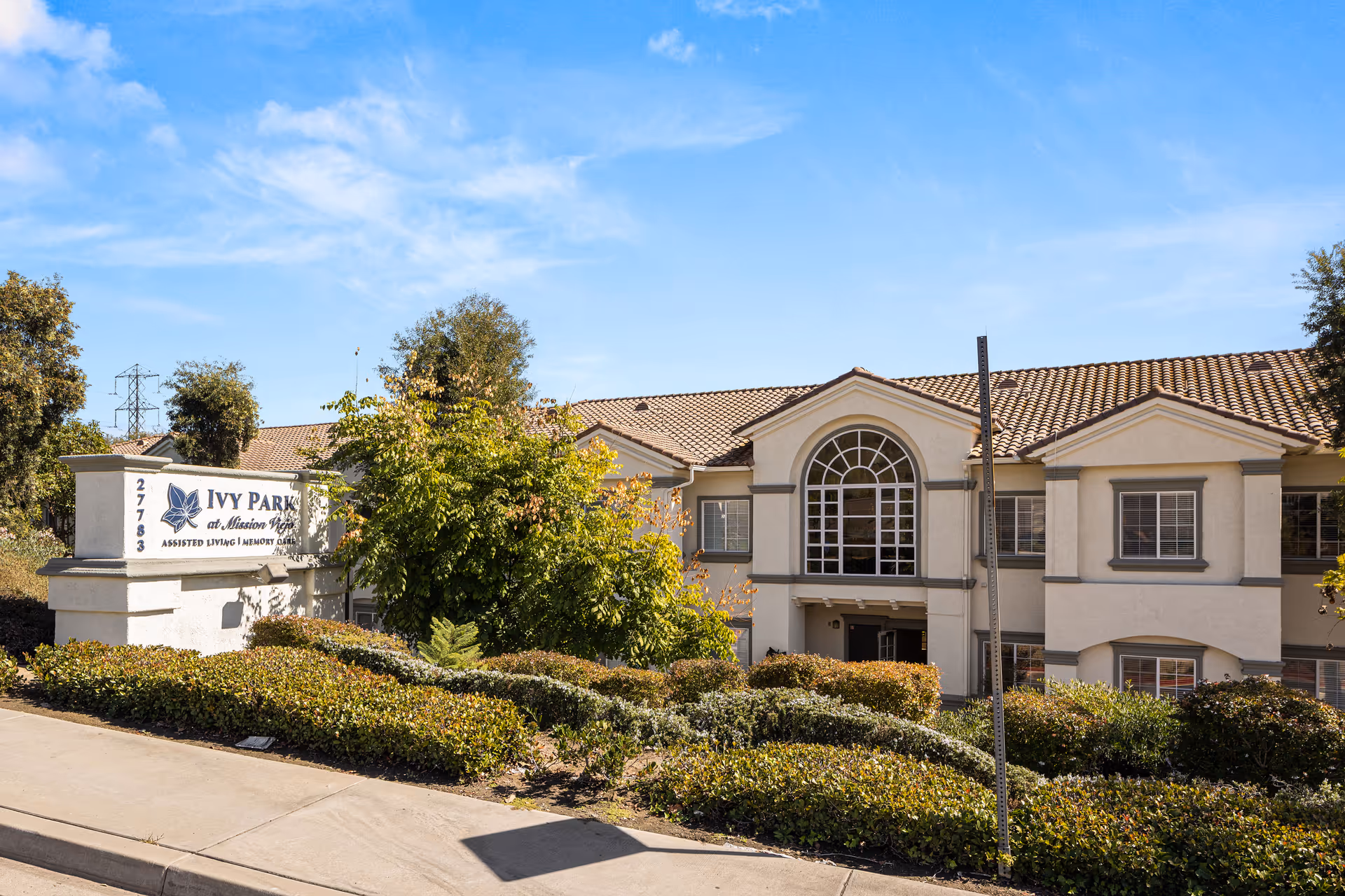 Exterior view of Ivy Park at Mission Viejo, a two-story assisted living and memory care facility with beige walls, a tiled roof, and large arched windows. The building is surrounded by green bushes and trees under a clear blue sky. A sign with the facility's name and address is visible on the left side near the sidewalk.