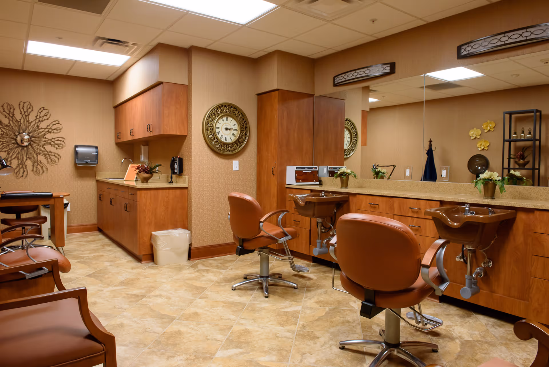 Interior view of a salon area in a senior living facility with two brown salon chairs in front of sinks and a large mirror. The room has wooden cabinets, a wall clock, decorative wall art, and a tiled floor.