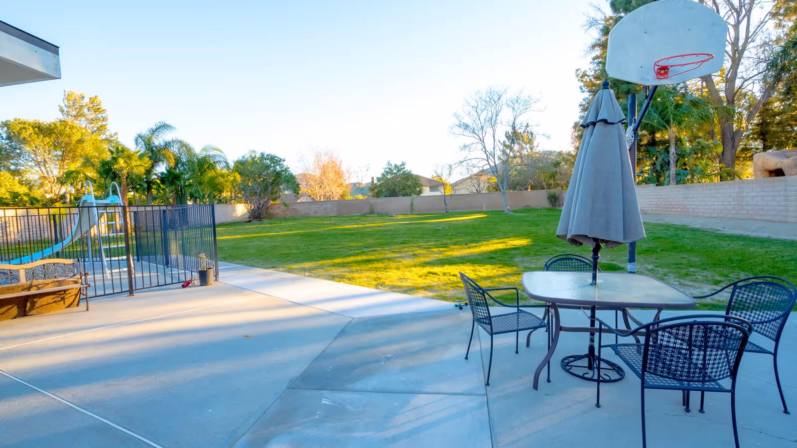 Outdoor patio area with a glass table and four metal chairs, a closed umbrella in the center of the table, a basketball hoop, a fenced swimming pool with a slide, and a large grassy yard surrounded by trees and a brick wall.