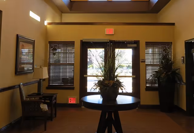 Interior view of an entrance area with double glass doors letting in natural light. A round table with a large floral arrangement is centered in front of the doors. There are two windows with wooden blinds on either side of the doors, a chair and side table with a lamp on the left, and a tall plant in a pot on the right. The walls are painted a warm beige color and there are exit signs above and below the doors.