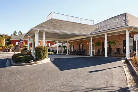 Exterior view of Cedarhurst of Oakwood showing a covered entrance with white pillars, rocking chairs along the brick wall, and an American flag near the driveway. The area is paved and landscaped with bushes and trees in the background under a clear sky.