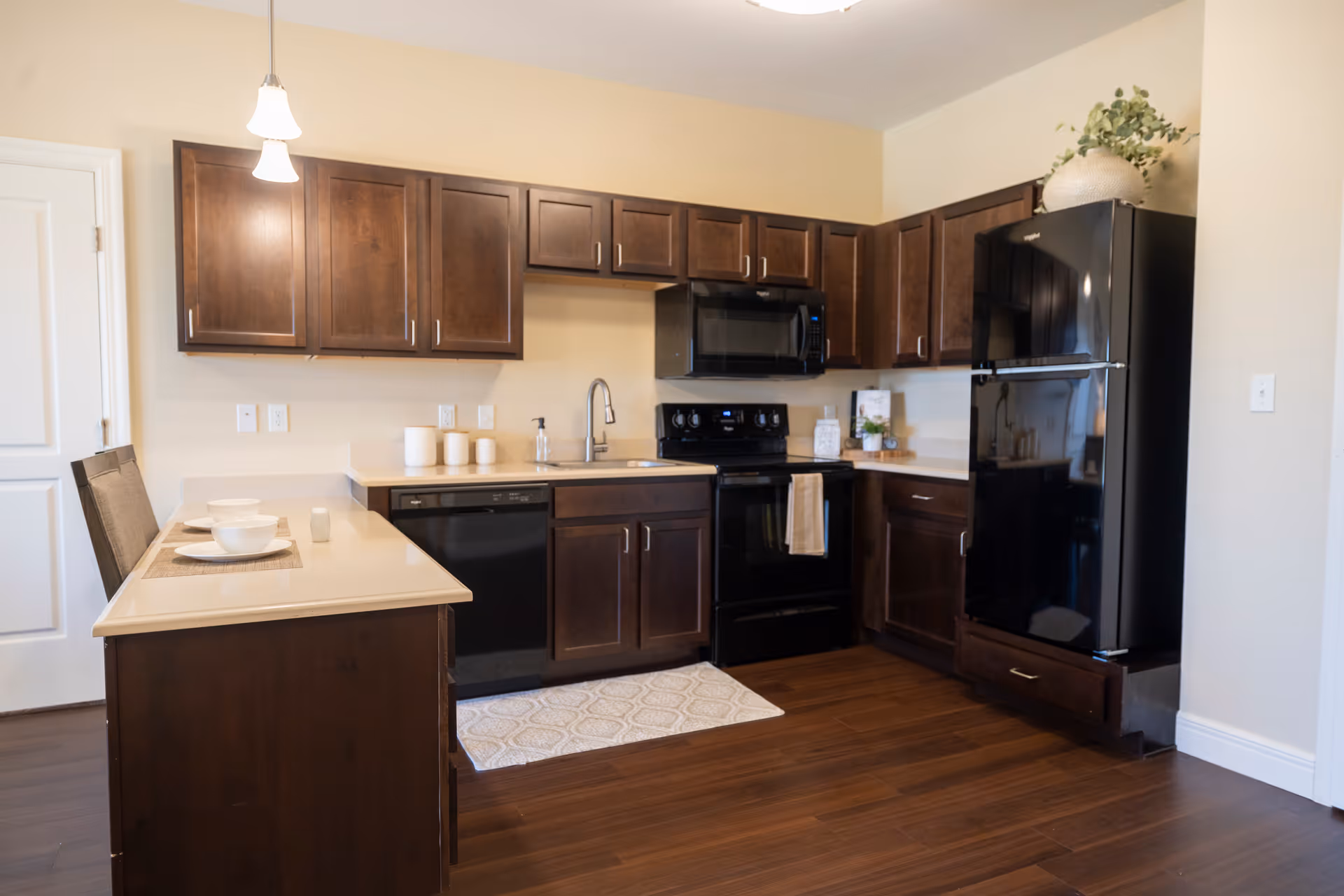 Modern kitchen with dark wood cabinets, black appliances including a refrigerator, microwave, stove, and dishwasher. There is a beige countertop with two place settings on a peninsula, a sink with a faucet, and a small rug on the wooden floor.