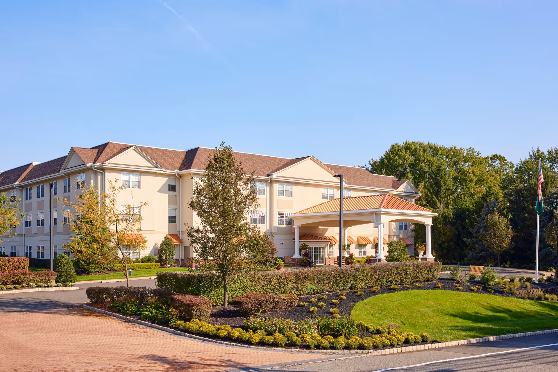 Front exterior of a three-story senior living facility with a porte-cochère entrance, landscaped grounds, and flagpoles.