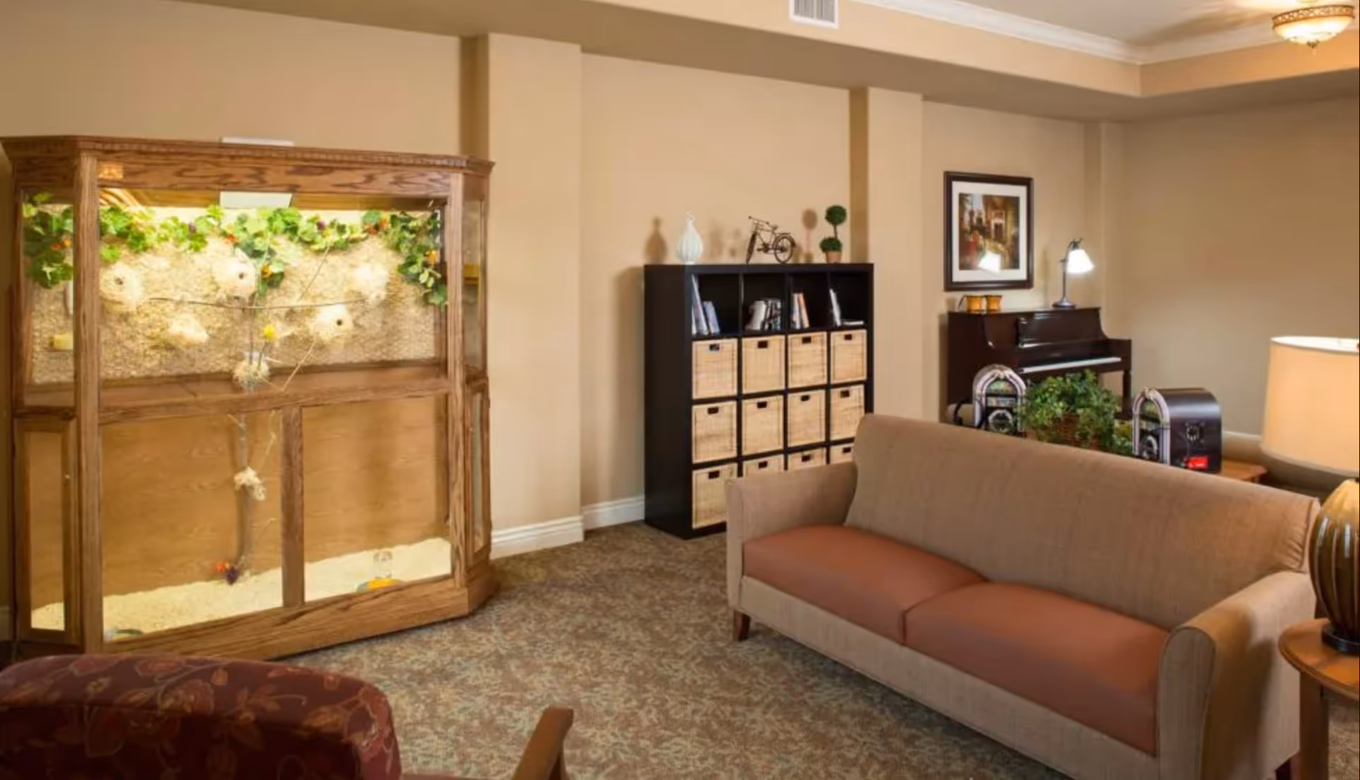 A cozy living room area with a beige sofa, a wooden display case with a nature-themed background, a black shelving unit with wicker baskets, a piano with a lamp and framed artwork above it, and a patterned carpeted floor.