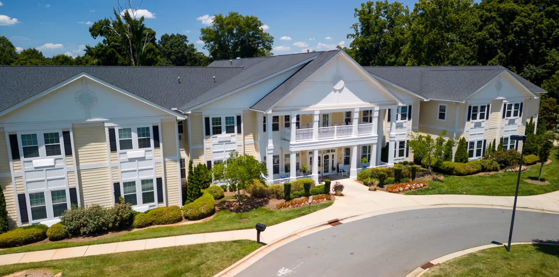 Exterior view of a large, two-story senior living facility building with beige siding, white trim, and a dark gray roof. The entrance features a covered porch with white columns and a balcony above. The surrounding area is landscaped with green bushes, trees, and flower beds, and a curved driveway leads to the entrance.