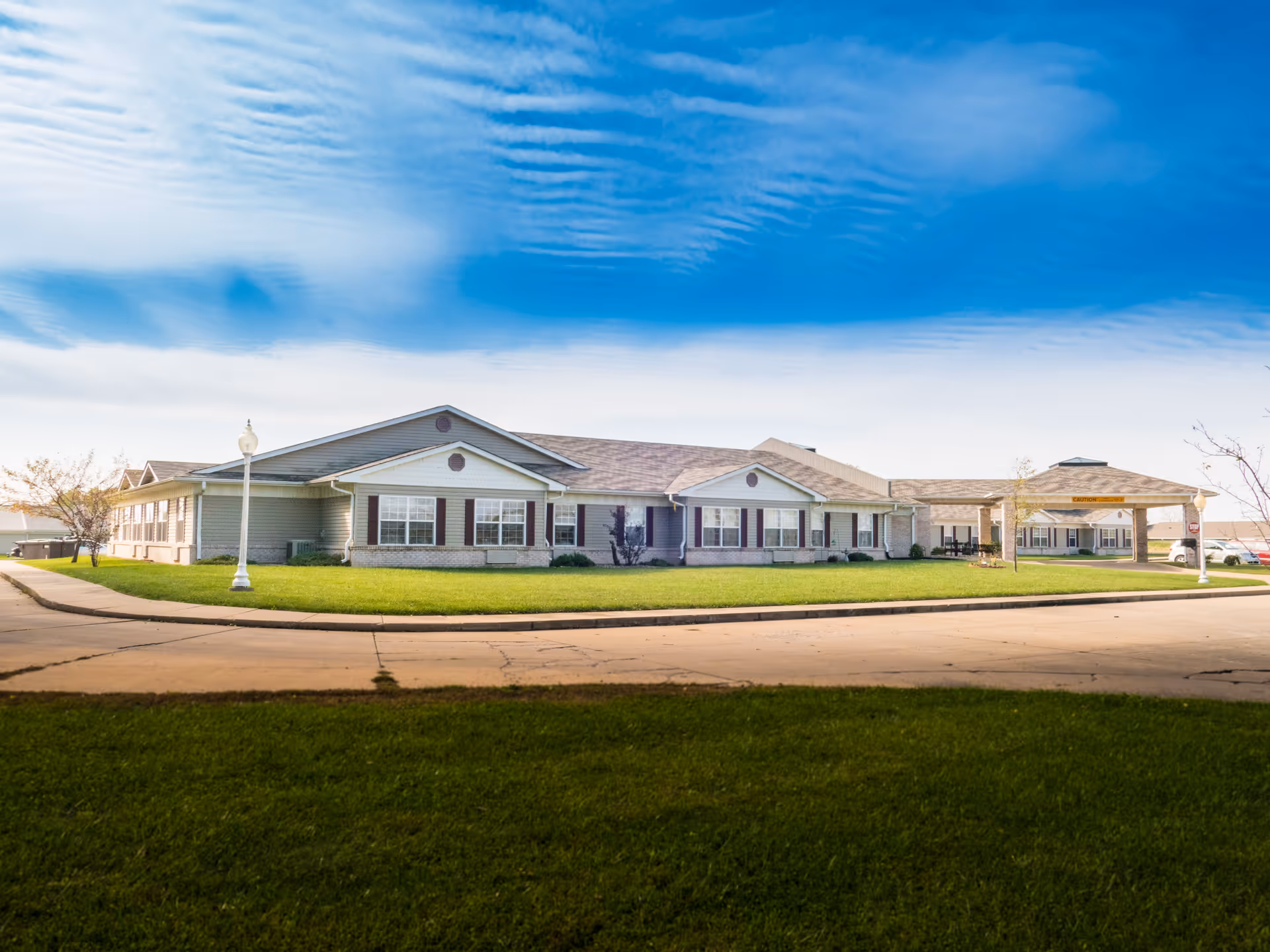 Exterior view of RidgeView Assisted Living, a single-story building with beige siding and brick accents, multiple windows with dark shutters, a covered entrance with a caution sign, surrounded by green grass and a paved driveway under a partly cloudy blue sky.