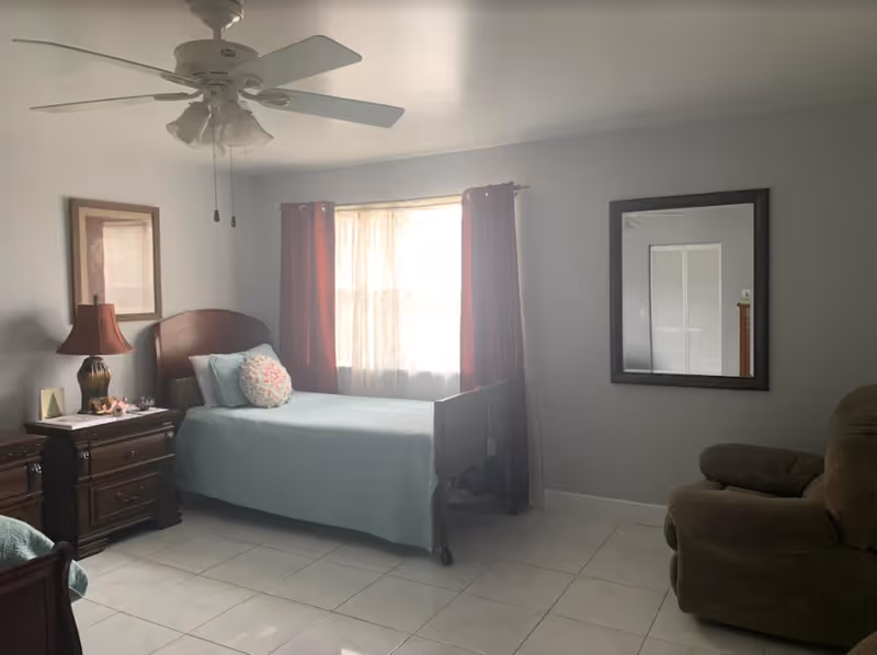 Bedroom in a senior living center with bed, nightstands, lamp, mirror, and window.
