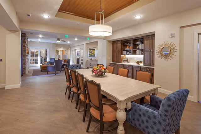 A spacious dining area with a long wooden table surrounded by eight chairs, including two blue upholstered armchairs at the ends. The ceiling above the table features a wooden inset with a modern hanging light fixture. In the background, there is a built-in wooden cabinet with shelves displaying decorative items and a sunburst mirror on the wall. Beyond the dining area, there is a lounge space with additional seating and a stone fireplace.