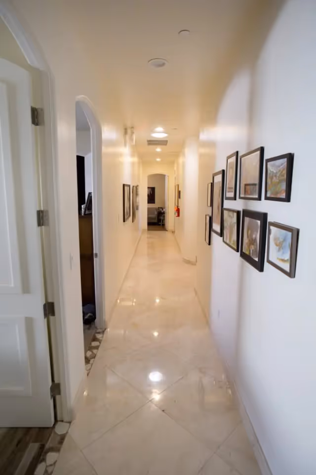 A long, narrow hallway with white walls and a shiny tiled floor. Several framed pictures hang on the right wall, and there are doorways on the left side. The hallway is well-lit with ceiling lights and leads to another room at the far end.