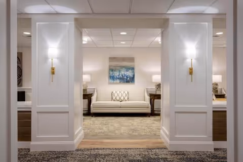 A well-lit interior space featuring a white cushioned sofa with patterned pillows centered between two wooden side tables with lamps. The walls are white with decorative paneling and two modern wall sconces. A framed abstract painting hangs above the sofa, and the floor is covered with a patterned carpet.