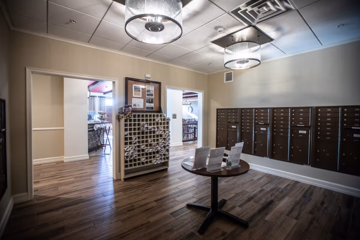 Interior view of a senior living facility mailroom with multiple mailboxes mounted on the wall, a wooden cubby shelf holding rolled-up papers, and a round table with informational pamphlets. The room has wood flooring, beige walls, and modern ceiling lights. Two doorways lead to other areas of the facility.