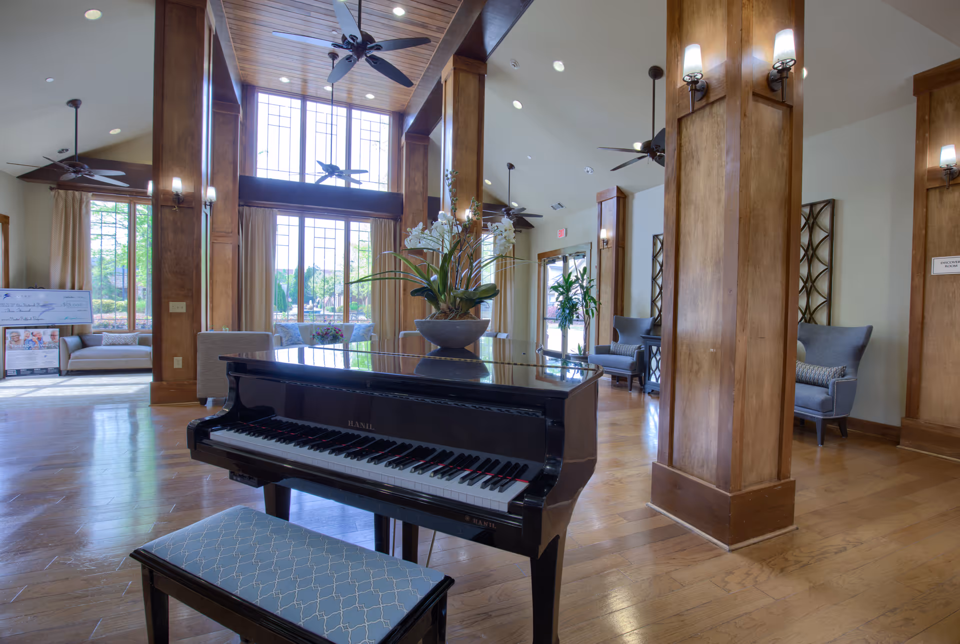 Spacious and well-lit common area with a black grand piano and a cushioned bench in the foreground. The room features large windows with natural light, wooden columns with wall sconces, ceiling fans, and comfortable seating areas with armchairs and sofas. A decorative plant is placed on top of the piano.