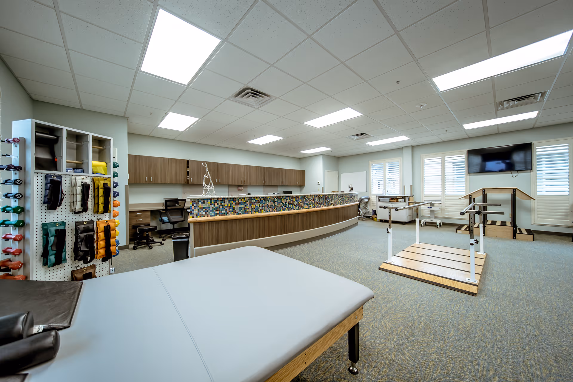 Bright physical therapy room with treatment tables, parallel bars, exercise equipment and a curved reception counter.