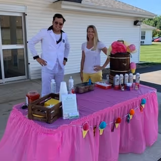 A man and a woman standing behind a table covered with a pink tablecloth decorated with colorful pom-poms and pennants. The table has various items including bottles, a wooden crate, and a barrel with pink cotton candy. They are outside near a white building on a sunny day.