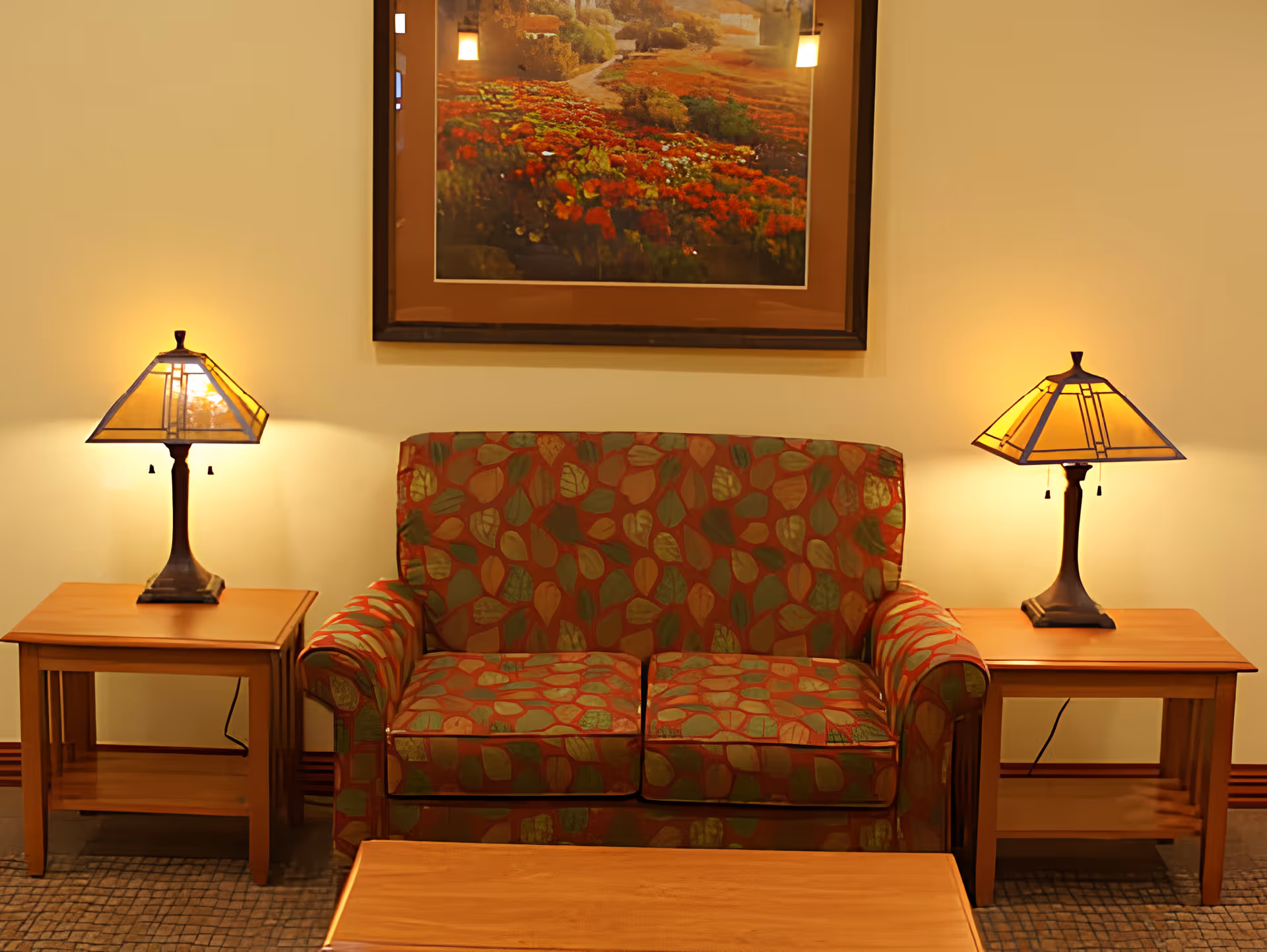 A cozy seating area with a patterned loveseat featuring red, green, and beige leaf designs, flanked by two wooden side tables each with a decorative table lamp emitting warm light. Above the loveseat hangs a framed picture of a vibrant flower garden.