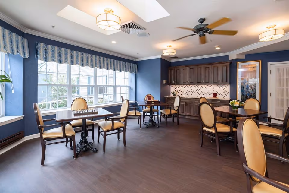 A bright and spacious common room with large windows letting in natural light. The room features several tables with beige cushioned chairs arranged around them. One table has a chessboard set up. The walls are painted blue with white trim, and there are dark wooden cabinets along one wall with a decorative backsplash. The ceiling has modern light fixtures and a ceiling fan. A framed artwork is visible on the right wall.