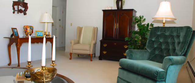 A cozy living room with a green upholstered armchair, a beige wingback chair, a dark wooden cabinet, a wooden side table with framed photos and a colorful vase, a glass coffee table with a brass candle holder and three white candles, and two table lamps providing warm lighting.