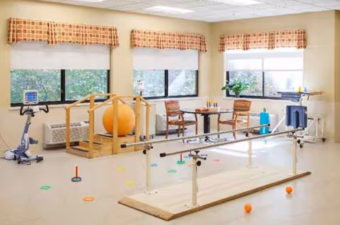 Interior rehabilitation gym with parallel bars, exercise balls, and chairs by windows.