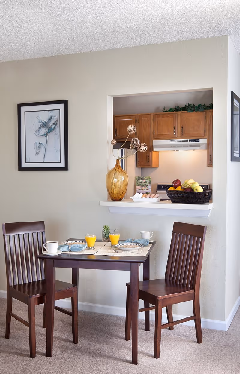 A small dining area with a dark wooden table and two matching chairs. The table is set with two plates, two glasses of orange juice, two cups, and napkins. Behind the table is a pass-through window to a kitchen with wooden cabinets, a stove, and a basket of fruit on the counter. A large amber vase with decorative branches sits on the pass-through ledge. A framed floral artwork hangs on the wall to the left.