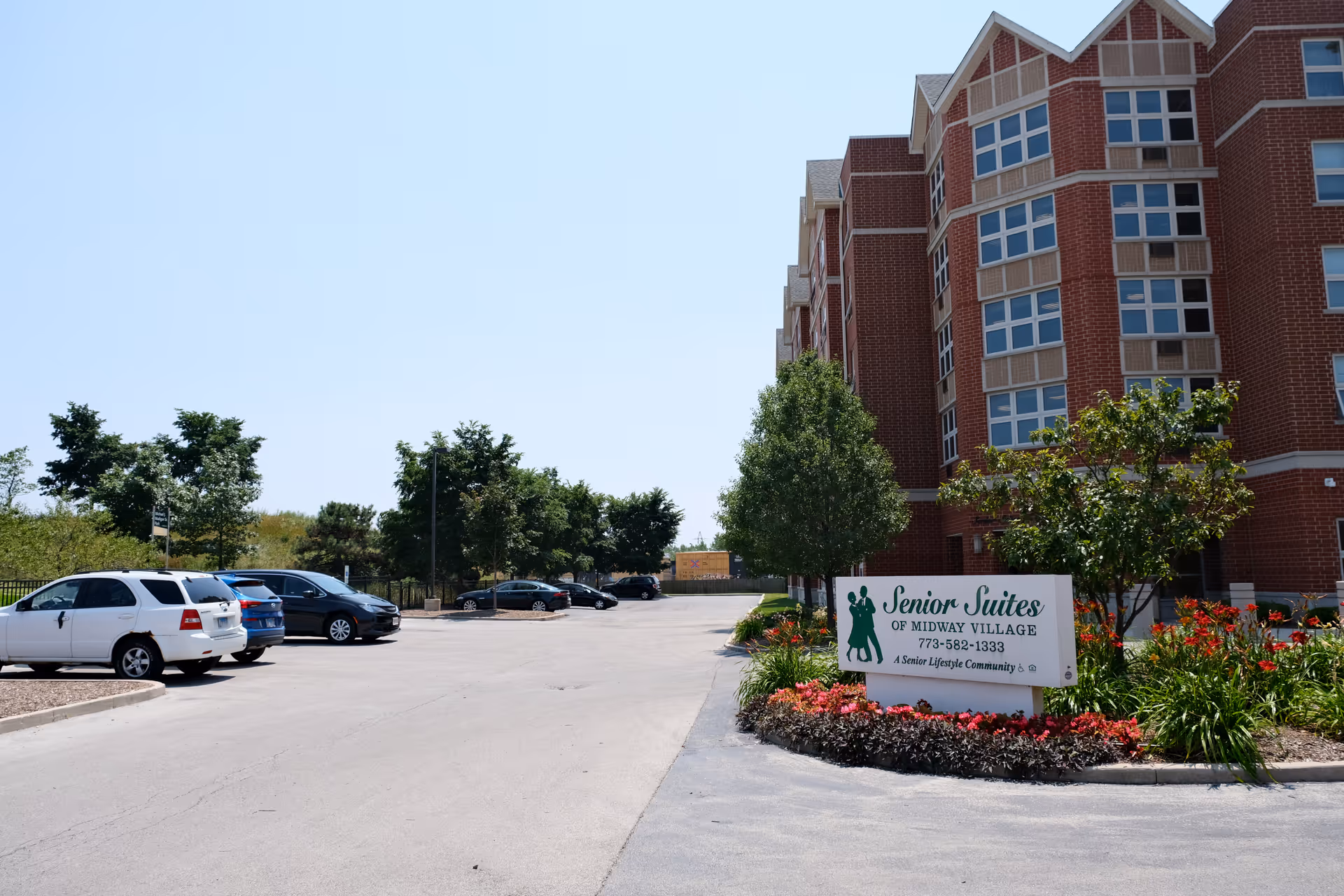 Exterior view of a senior living facility named Senior Suites of Midway Village, showing a multi-story brick building with many windows, a parking lot with several parked cars, and a landscaped area with flowers and trees near a sign displaying the facility's name and contact information.