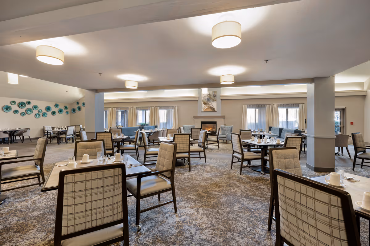 A spacious dining room in a senior living community with multiple tables and chairs arranged neatly. The room features soft lighting from ceiling fixtures, a carpeted floor, large windows with curtains, and a fireplace with armchairs around it. Decorative wall art is visible on one side.
