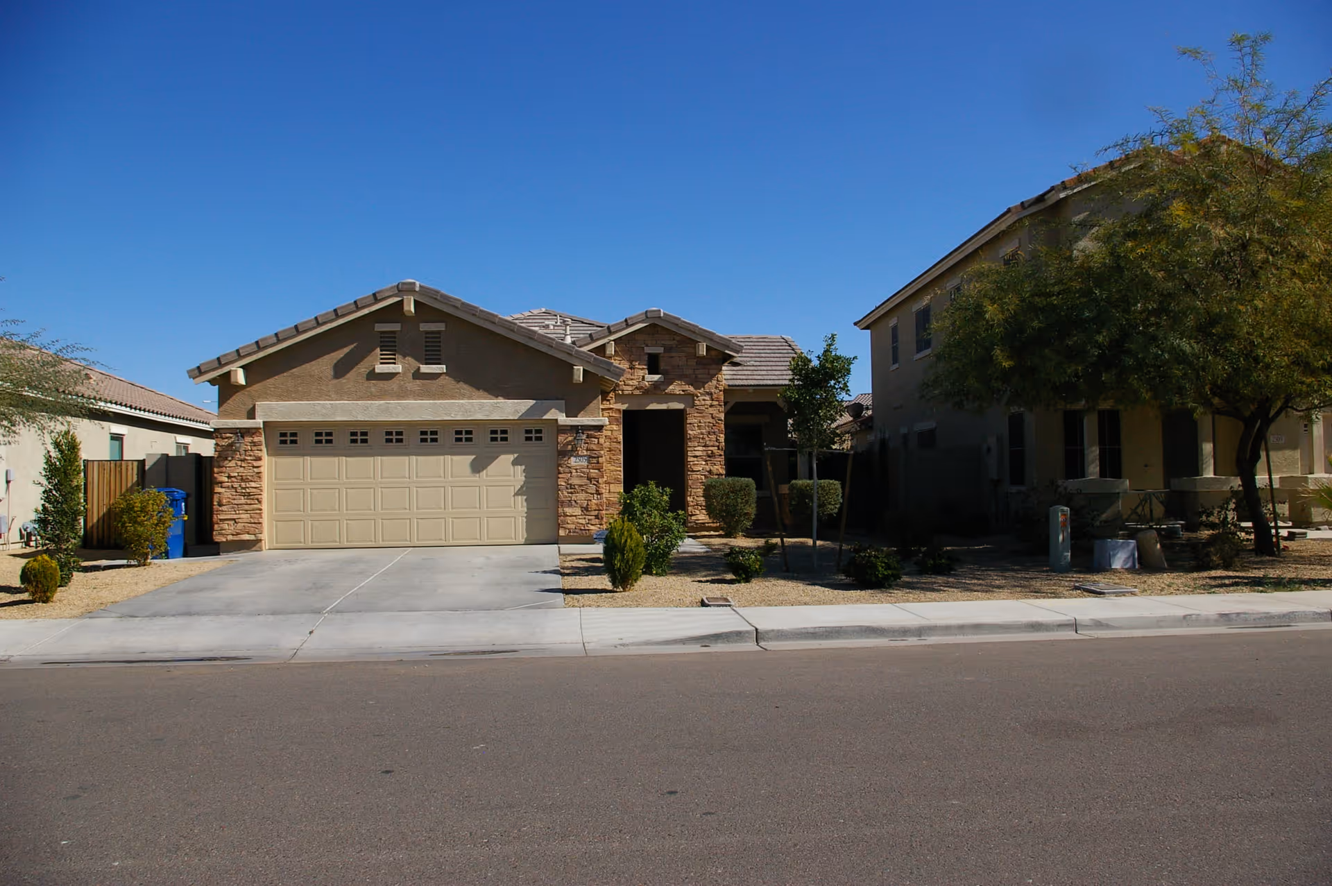 A single-story house with a beige exterior and stone accents, featuring a two-car garage and a driveway. The front yard has desert landscaping with small bushes and trees. The sky is clear and blue.