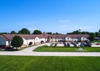 Aerial view of a single-story senior living facility building with a red roof and beige walls, surrounded by a green lawn and a parking lot with several cars. Trees and a water tower are visible in the background under a clear blue sky.