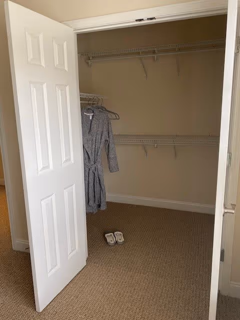 Open closet with white double doors, beige walls, and carpeted floor. Inside the closet, there are two wire shelves and a gray bathrobe hanging on a hanger. A pair of white slippers is placed on the floor in front of the closet.
