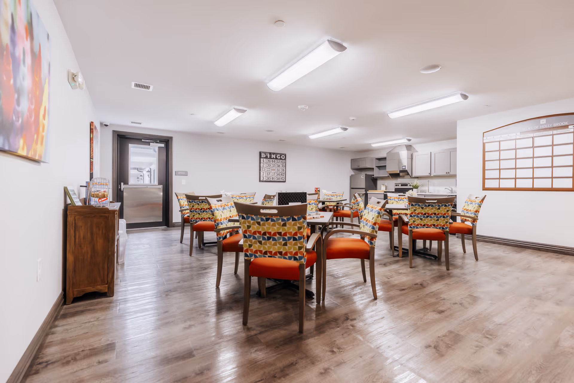 A bright, spacious room with multiple tables and colorful patterned chairs with orange seats arranged for group activities or dining. The room has a wood floor, white walls, and fluorescent ceiling lights. There is a kitchen area with cabinets, a stove, and a refrigerator in the background. A bingo board is mounted on the far wall, and a large whiteboard or schedule board is on the right wall. A wooden cabinet with some items on top is on the left side near the entrance door.