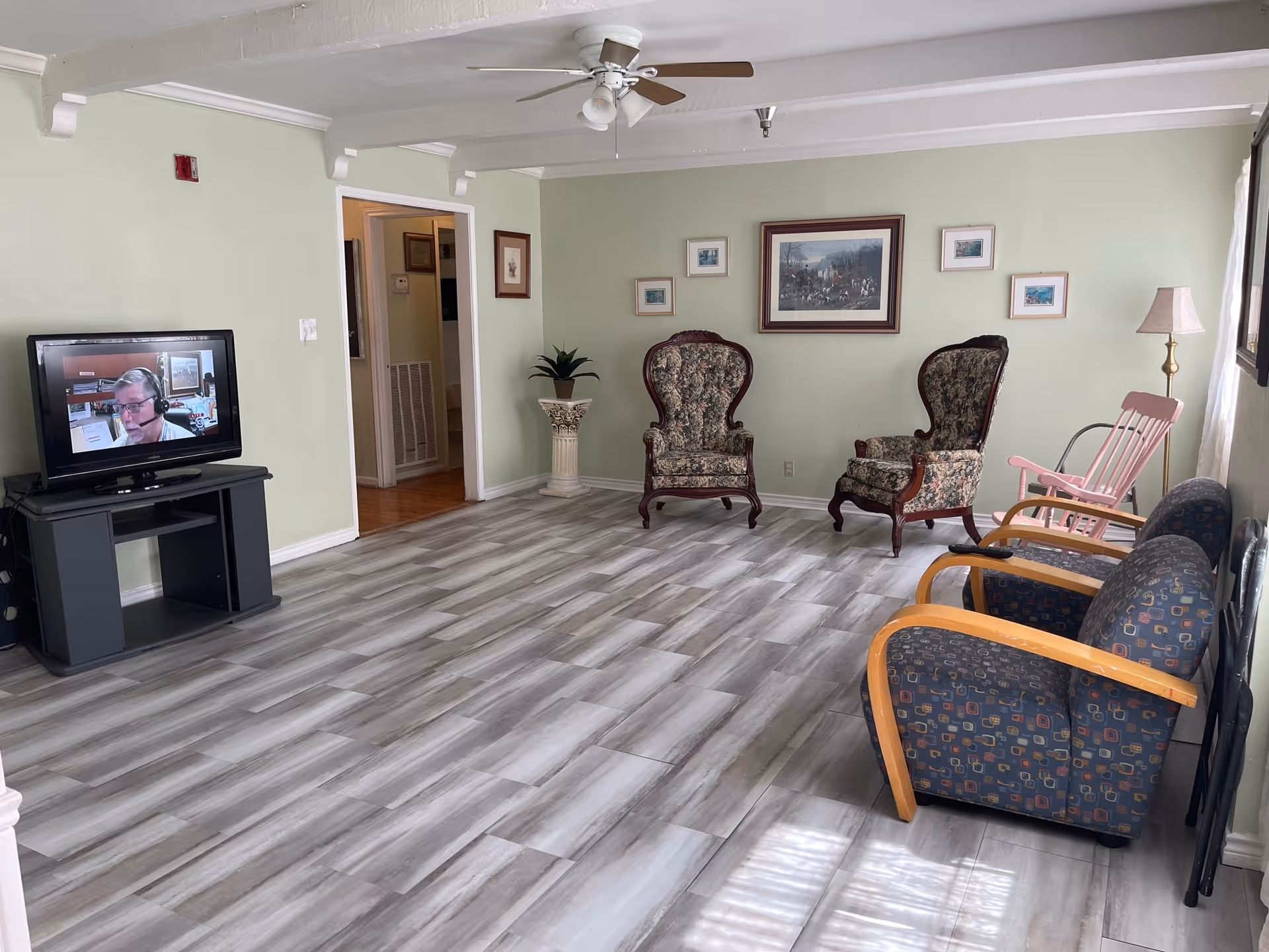 A bright common living room with a TV on a stand, patterned armchairs and rocking chairs along pale green walls and gray wood-look tile flooring.