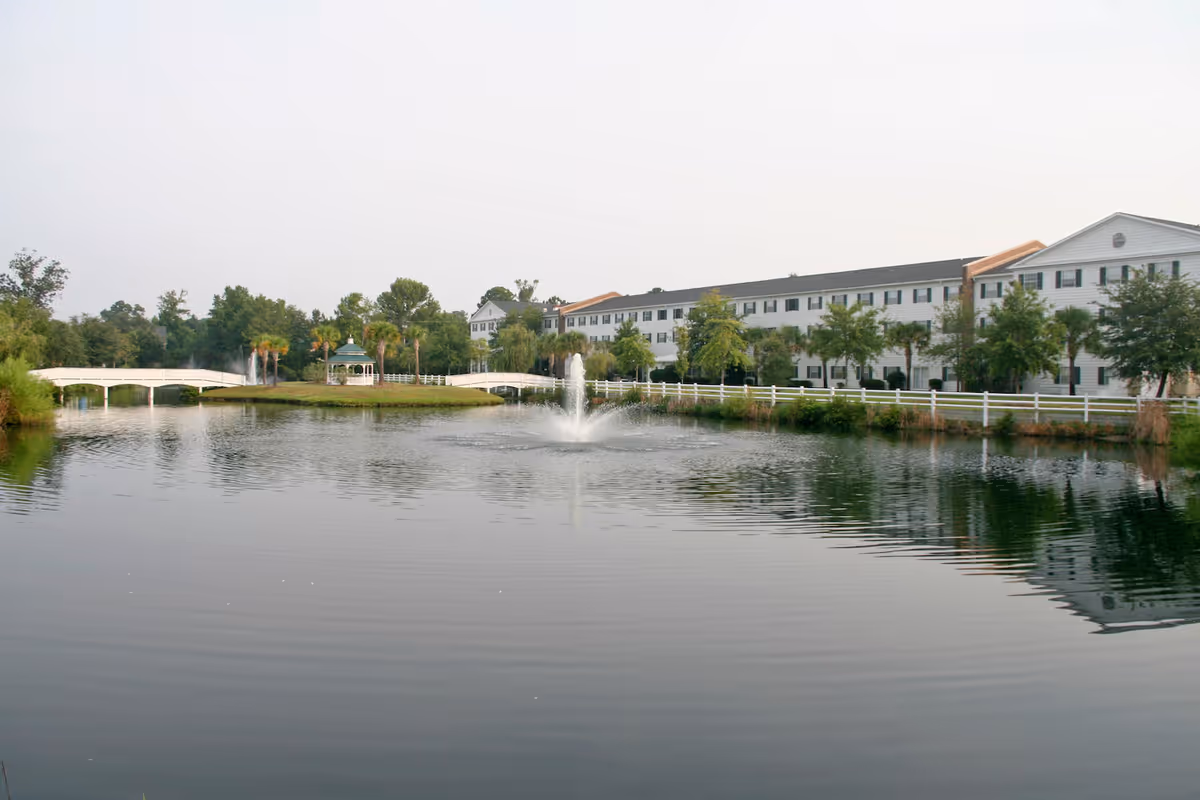 A large pond with a water fountain in the center, surrounded by greenery and trees. In the background, there is a long white building with multiple windows and a white fence running along the edge of the pond. A white bridge and a small gazebo are visible on the left side of the pond.