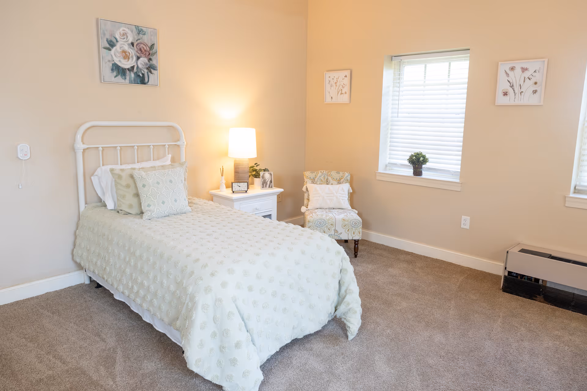 A cozy bedroom with a single bed covered in a light green textured bedspread and matching pillows. Next to the bed is a white nightstand with a lamp, a small plant, a clock, and a framed photo. A patterned armchair with a white pillow is placed near two windows with white blinds. The walls are painted beige and decorated with floral artwork. The floor is carpeted in a neutral tone.