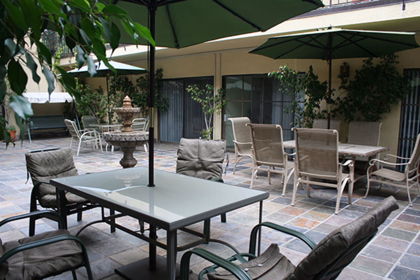 Outdoor patio area with multiple tables and chairs, some with green umbrellas providing shade. There is a decorative stone fountain in the center and potted plants along the walls of the building.