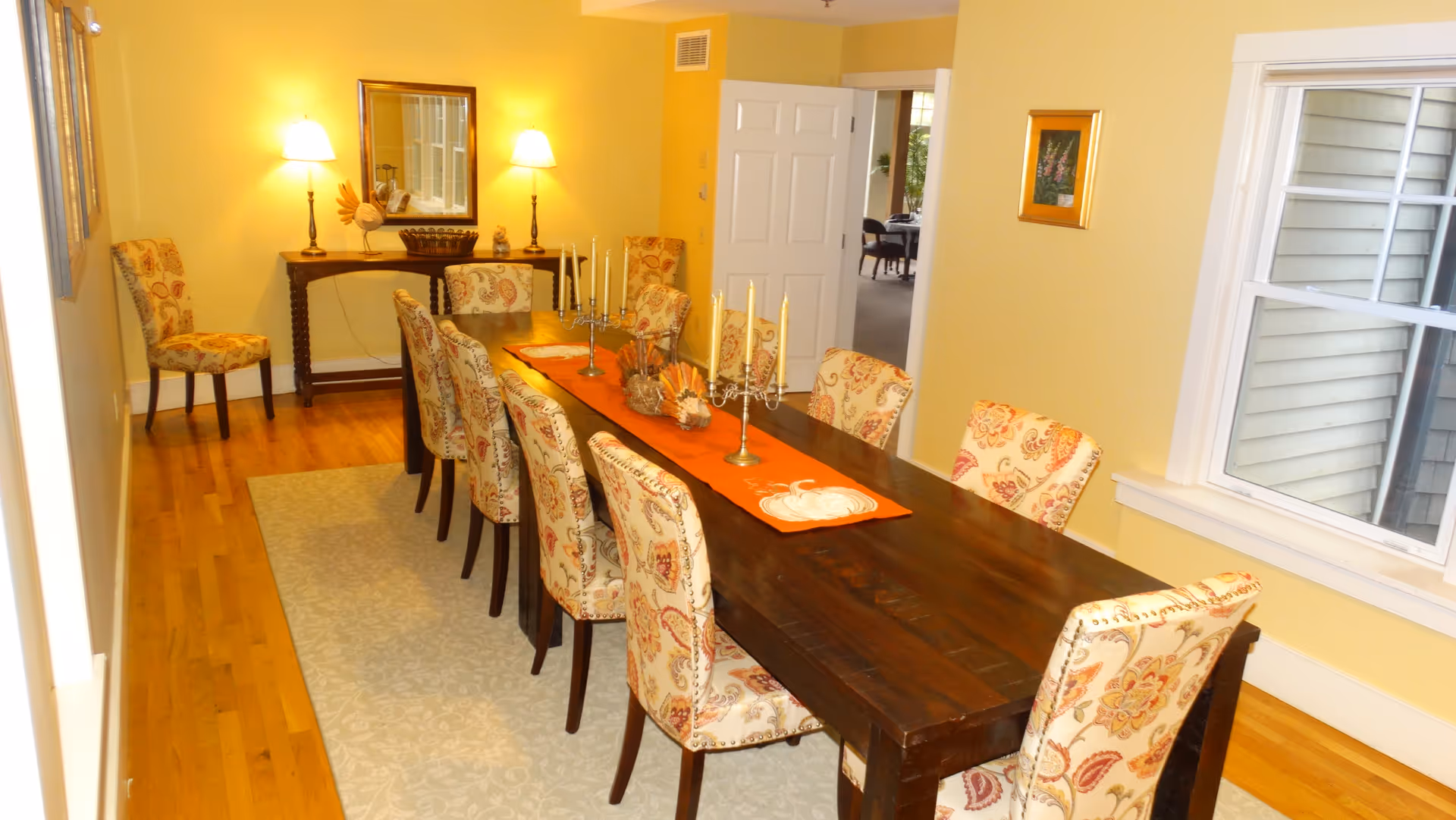 A dining room with a long dark wooden table surrounded by ten upholstered chairs with floral patterns. The table is decorated with an orange runner, candle holders, and small decorative items. The room has yellow walls, hardwood floors, a light-colored rug under the table, a window on the right, and a console table with two lamps and a mirror on the far wall.