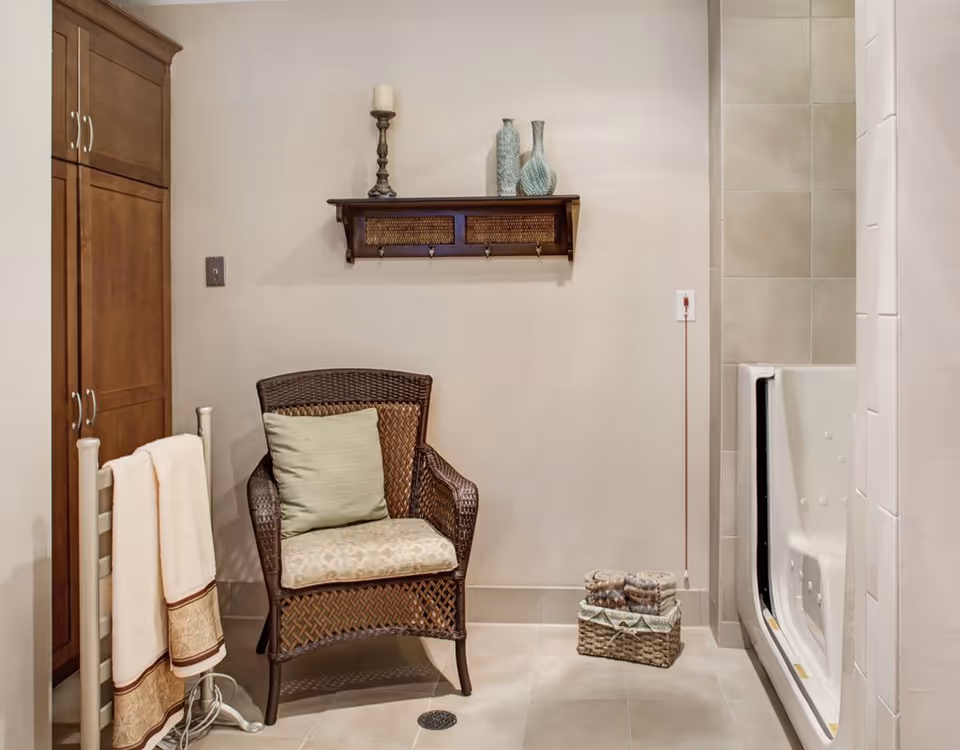 A bathroom area featuring a wicker chair with a cushion and pillow, a towel rack with two towels, a wooden cabinet, a wall shelf with decorative vases and a candle, and a walk-in bathtub with grab bars and jets.
