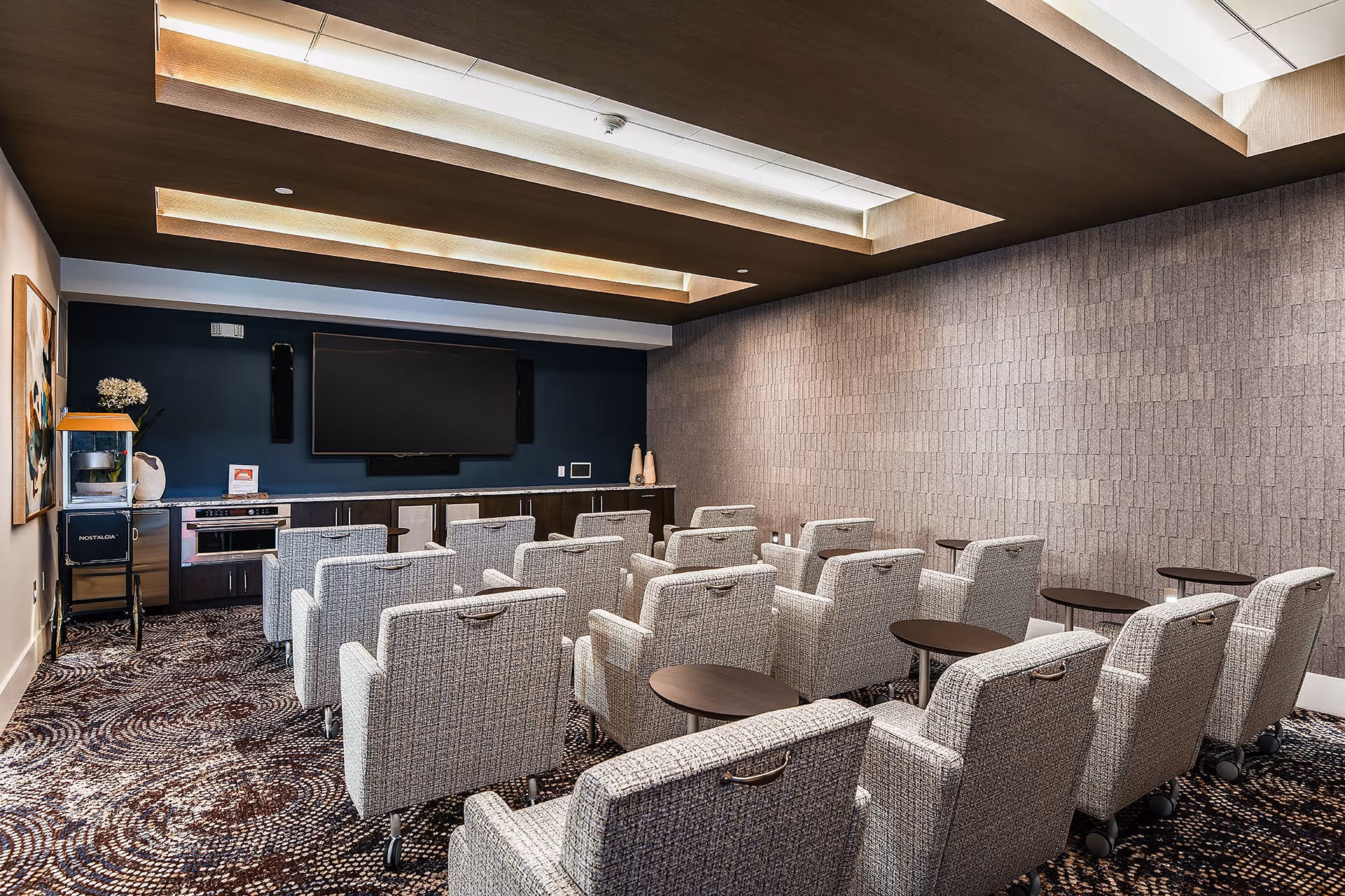 A small theater-style room with multiple rows of cushioned chairs facing a large flat-screen TV mounted on a dark blue wall. The room has a patterned carpet, a popcorn machine on the left side, and a cabinet with decorative items beneath the TV. The ceiling features recessed lighting with wooden accents.