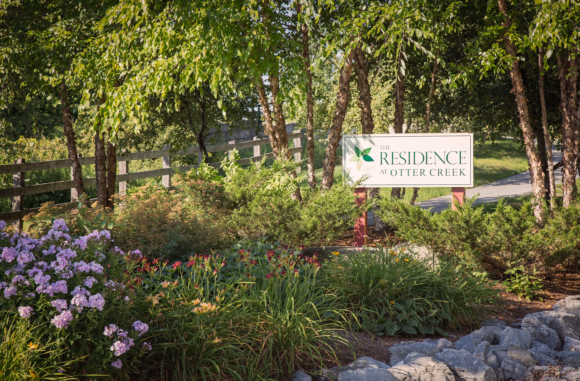A landscaped garden area with various green shrubs, flowering plants, and trees. A wooden fence runs along the left side, and a sign reading 'The Residence at Otter Creek' is prominently displayed among the greenery. A paved pathway is visible in the background.