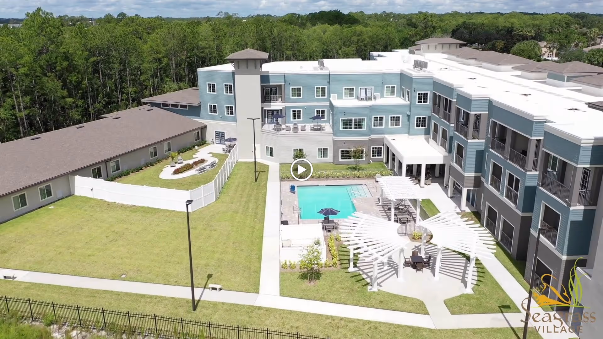 Aerial view of Seagrass Village of Port Orange senior living facility showing a large multi-story building with balconies, a swimming pool, a white pergola with seating underneath, a fenced garden area with benches, and surrounding green lawns with trees in the background.