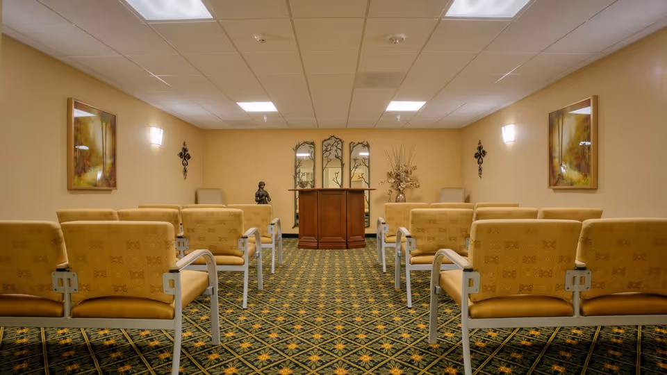 A small chapel or meditation room with rows of beige cushioned chairs facing a wooden podium. The room has patterned carpet, beige walls with framed artwork, wall sconces, and decorative items including a statue and a vase with dried flowers near the podium.