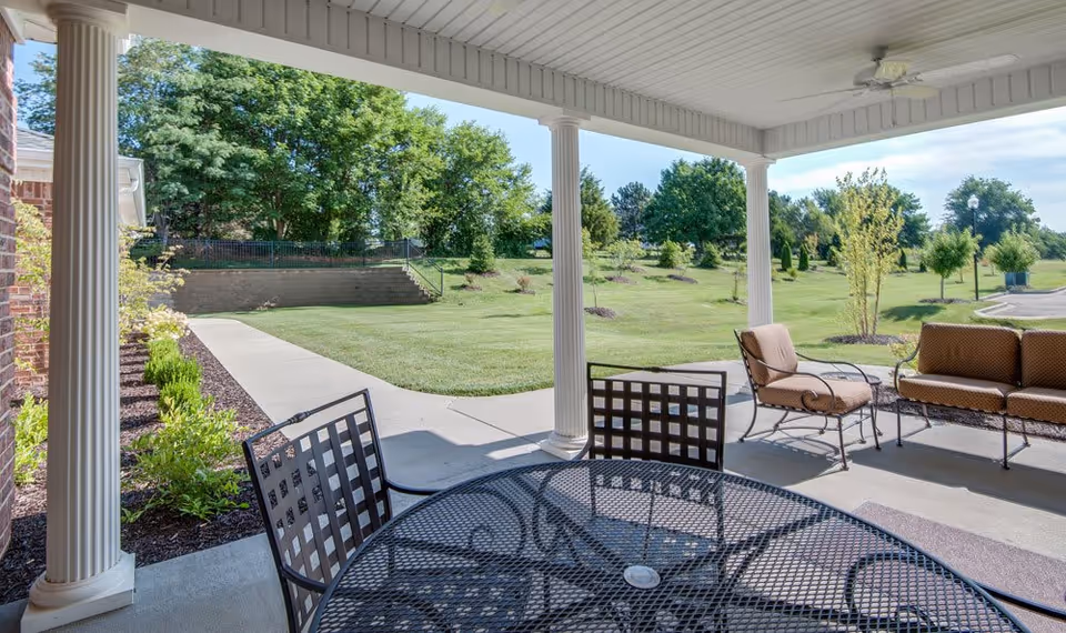 Covered patio with a metal table and chairs and cushioned seating overlooking a landscaped lawn and trees.