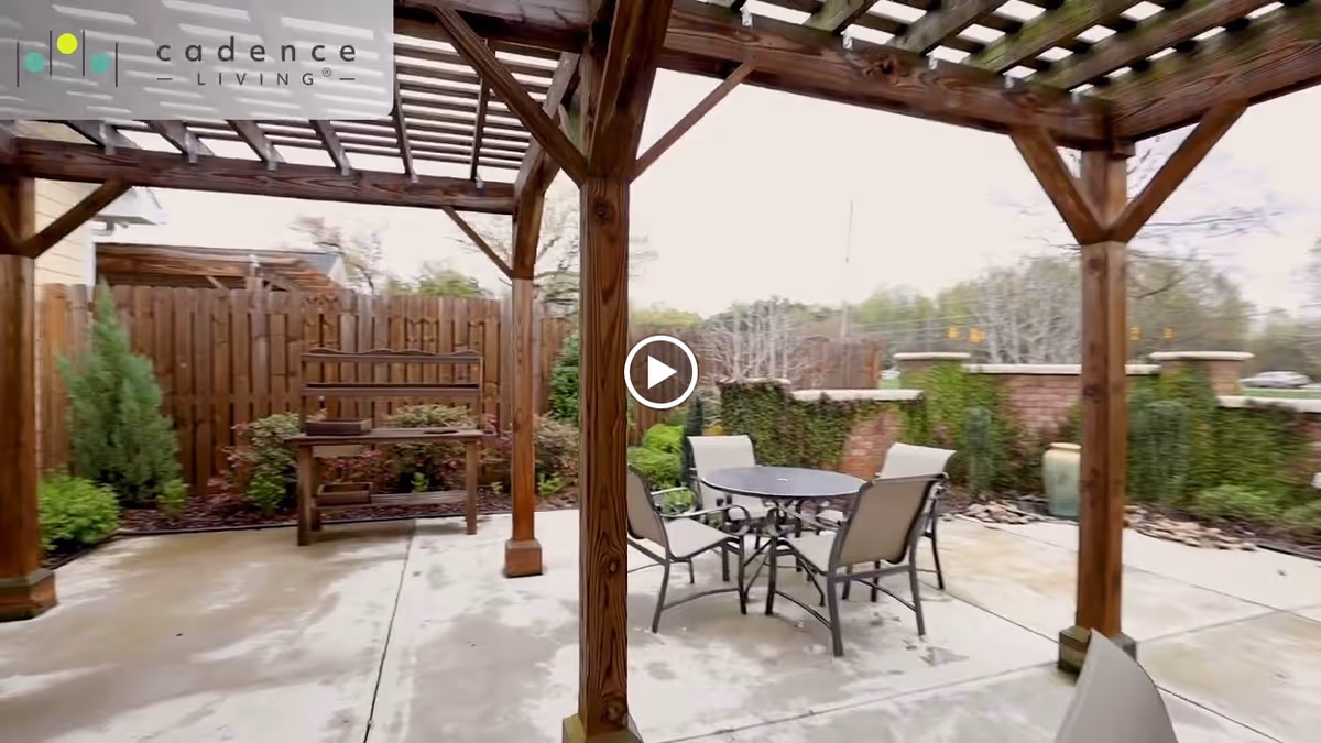 Outdoor patio area with a wooden pergola overhead, a round metal table with four chairs, a wooden bench against a wooden fence, and greenery including bushes and climbing plants on a brick wall.