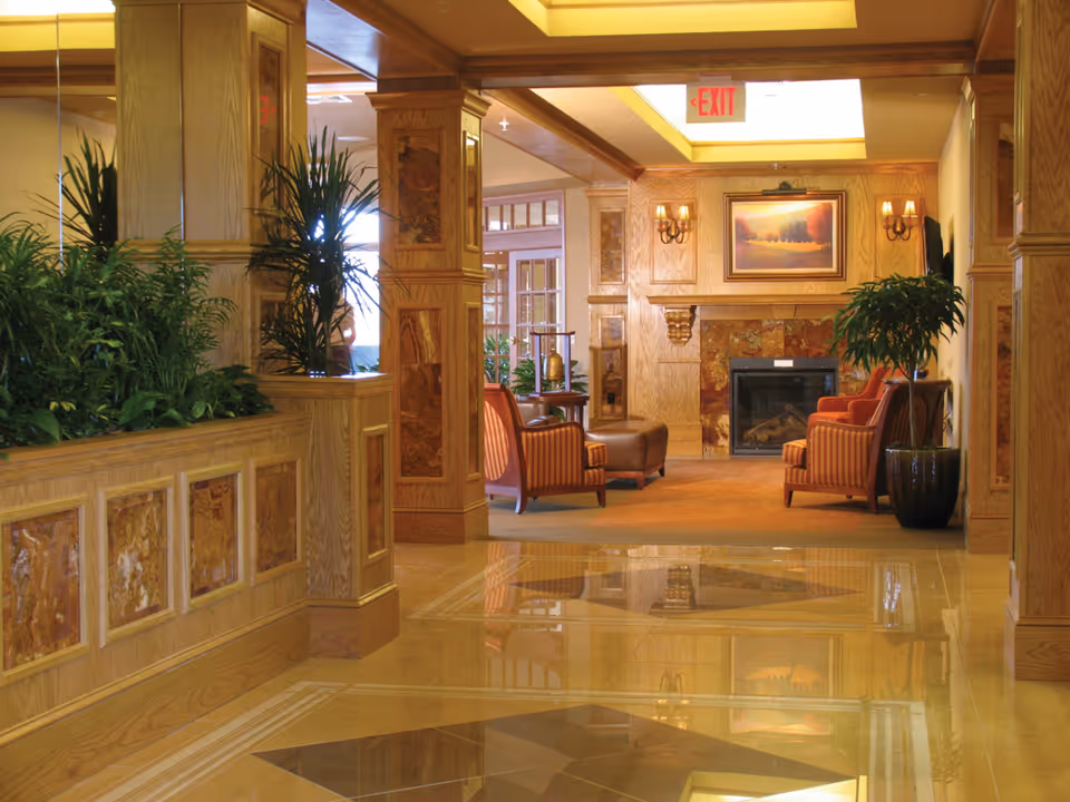 Wood-paneled senior living lobby with upholstered chairs, a fireplace, potted plants, and a reflective tiled floor.
