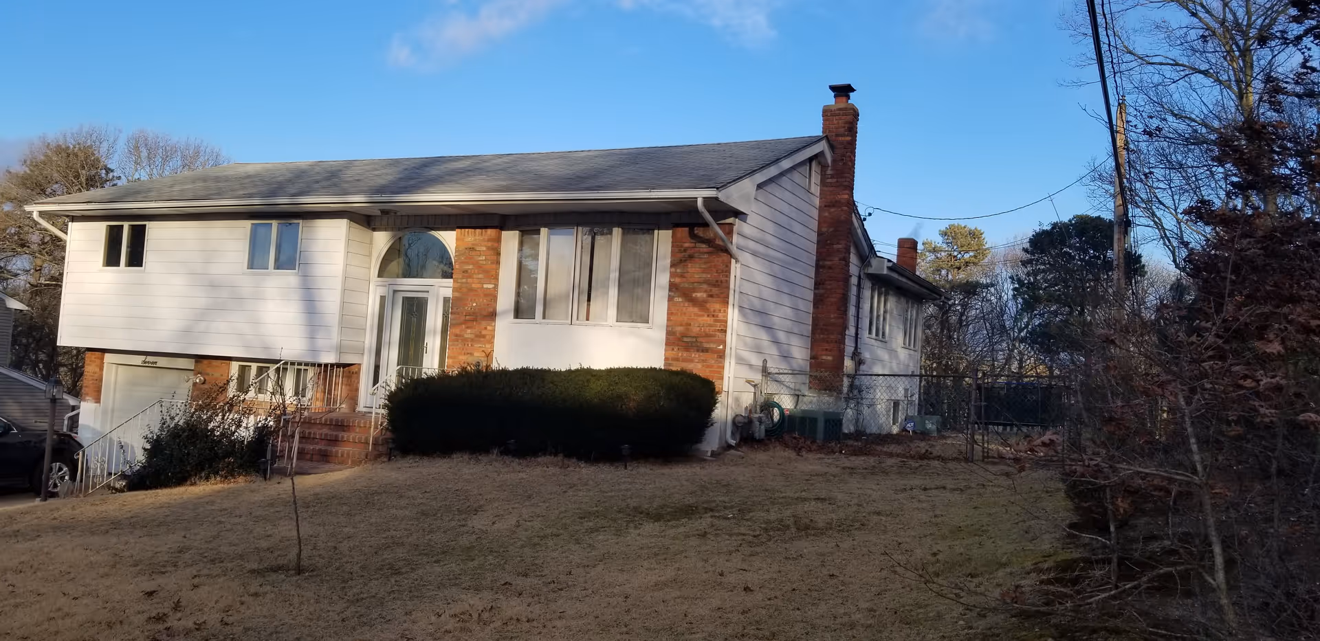 A single-story residential house with white siding and brick accents, a chimney, and a front door with an arched window above it. The house is surrounded by a lawn with some bushes and trees, under a clear blue sky.