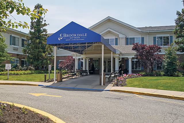 Entrance of Brookdale West Orange senior living facility with a blue canopy displaying the facility name. The building is two stories with white siding and surrounded by landscaped greenery and flowers. There are benches along the walkway leading to the entrance.
