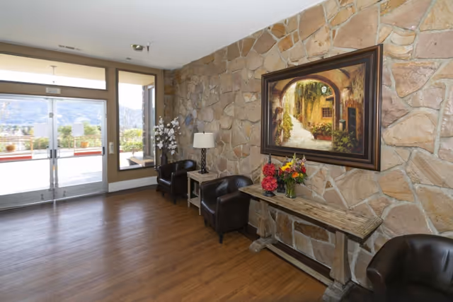 An interior seating area with a stone wall featuring a large framed painting of a sunlit archway and pathway. Below the painting is a rustic wooden console table with two vases of colorful flowers. Two dark leather chairs flank a small side table with a lamp and a decorative white flower arrangement. The floor is wooden, and there is a glass door and window letting in natural light, showing an outdoor view.