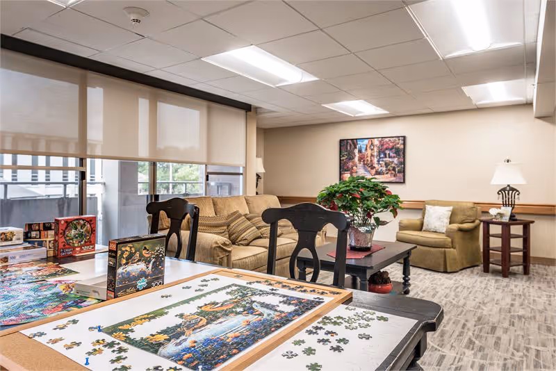 Communal activity room with a partially completed jigsaw puzzle on a table, sofas and armchairs, and a potted plant.
